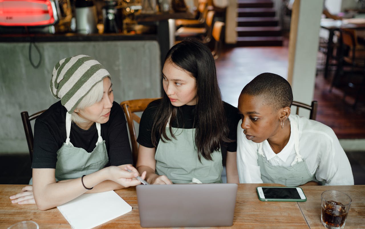 Three women in aprons deeply engaged in teamwork at a café table with a laptop.